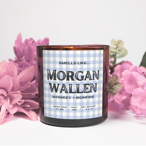 May include: A brown glass candle jar with a label that reads "Morgan Wallen Whiskey + Bonfire." The label has a blue and white gingham pattern. The candle is surrounded by pink flowers. The candle's net weight is 9.5 oz.
