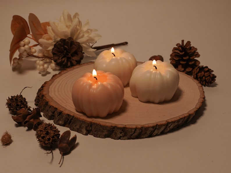 May include: Three pumpkin-shaped candles on a wooden slice. Two candles are white and one is peach. All candles are lit.