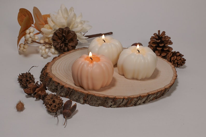 May include: Three pumpkin-shaped candles on a wooden slice. The candles are lit and are in shades of white and orange.