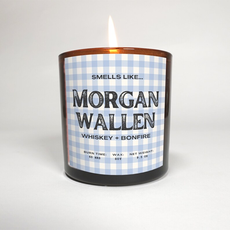 May include: A lit candle in a brown glass jar. The label features a blue and white gingham pattern and text that reads "Smells Like... Morgan Wallen Whiskey + Bonfire." The candle weighs 9.5 oz and has a burn time of 60 hours.