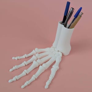 Op de afbeelding: Een witte, 3D-geprinte pennenhouder in de vorm van een menselijke skelet hand. De houder bevat pennen met blauwe, zwarte en zilveren schachten. De achtergrond is effen lichtroze. Een uniek bureau-accessoire.