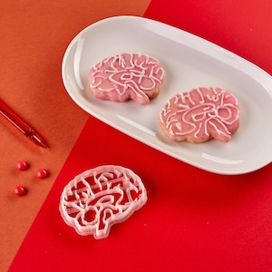 Puede incluir: Dos galletas de azúcar rosadas decoradas con glaseado blanco en forma de cerebro en un plato ovalado blanco. Tres corazones de caramelo rojos están en una superficie roja junto al plato.