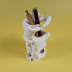 May include: White ceramic pen holder shaped like a human spine with two pens inside. One pen is brown wood and the other is black and silver.