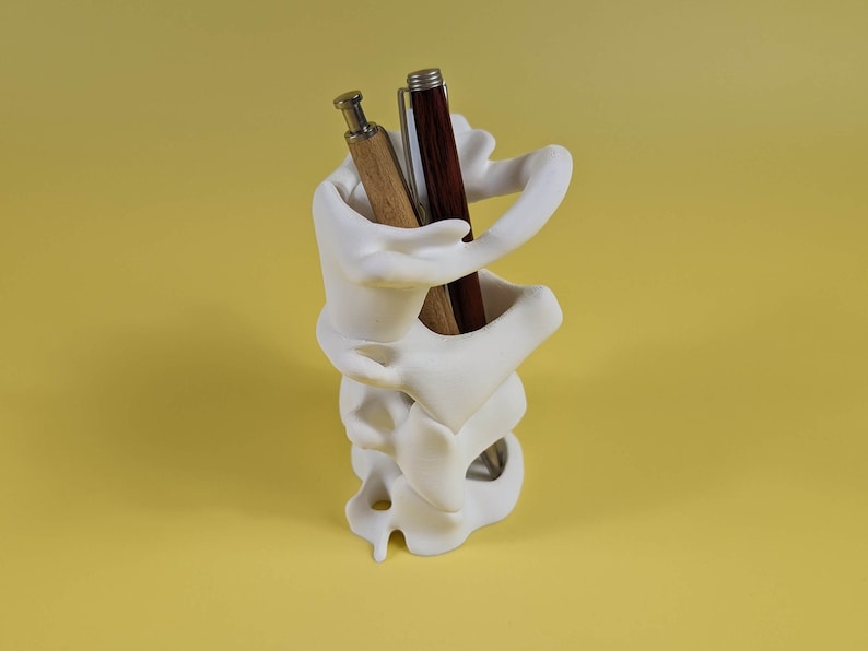 May include: White ceramic pen holder shaped like a human spine with two pens inside. One pen is brown wood and the other is black and silver.
