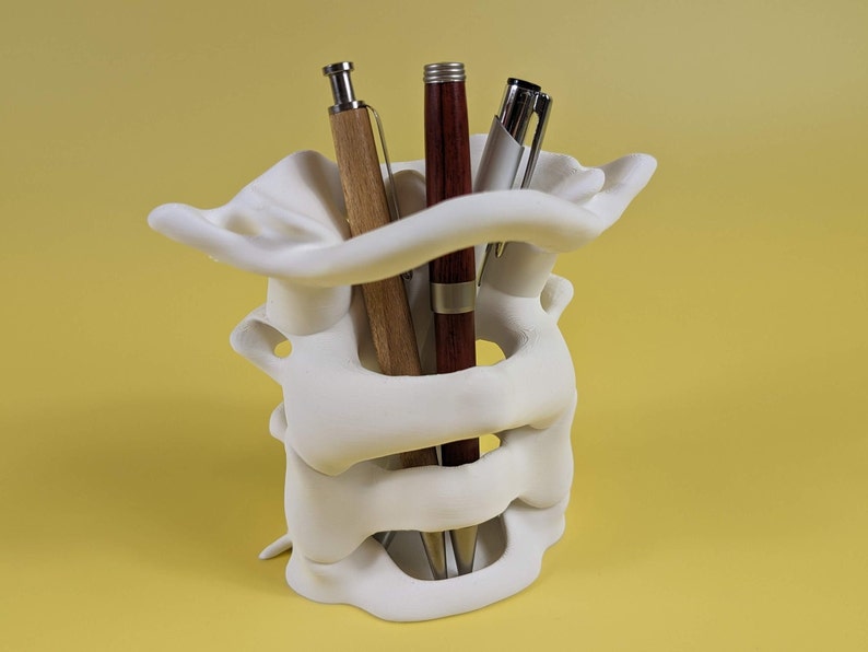 May include: White ceramic pen holder shaped like a human spine with three pens inside. The pens are brown, red, and silver.