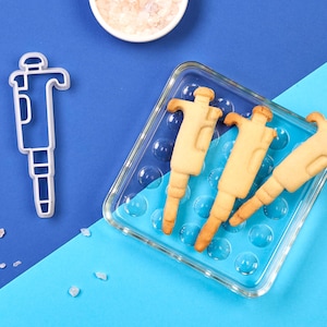 Può includere: Biscotti a forma di pipetta, presentati su un vassoio blu all'interno di un contenitore di vetro trasparente. È presente anche un tagliabiscotti a forma di pipetta. Lo sfondo è una sfumatura di blu, con una piccola ciotola di sale rosa.
