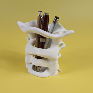 May include: White ceramic pen holder shaped like a human spine with three pens inside. The pens are brown, red, and silver.