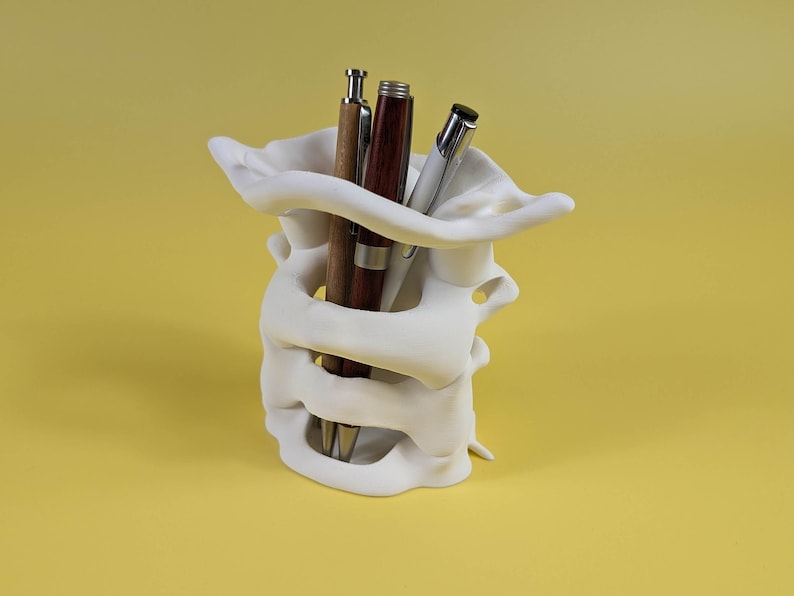 May include: White ceramic pen holder shaped like a human spine with three pens inside. The pens are brown, red, and silver.