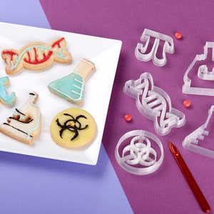 Puede incluir: Un plato blanco con galletas con tem&aacute;tica cient&iacute;fica decoradas con glaseado. Las galletas tienen forma de cadena de ADN, un vaso de precipitados, un microscopio, un s&iacute;mbolo de pastel y un s&iacute;mbolo de peligro biol&oacute;gico. Tambi&eacute;n hay cortadores de galletas de pl&aacute;stico transparente con forma de cadena de ADN, un vaso de precipitados, un microscopio, un s&iacute;mbolo de pastel y un s&iacute;mbolo de peligro biol&oacute;gico.