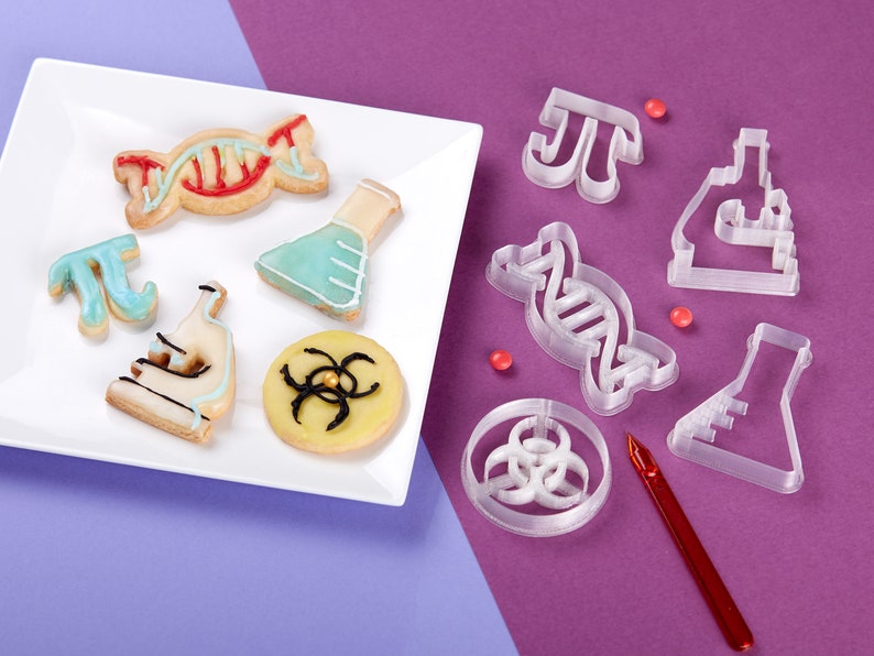 Puede incluir: Un plato blanco con galletas con tem&aacute;tica cient&iacute;fica decoradas con glaseado. Las galletas tienen forma de cadena de ADN, un vaso de precipitados, un microscopio, un s&iacute;mbolo de pastel y un s&iacute;mbolo de peligro biol&oacute;gico. Tambi&eacute;n hay cortadores de galletas de pl&aacute;stico transparente con forma de cadena de ADN, un vaso de precipitados, un microscopio, un s&iacute;mbolo de pastel y un s&iacute;mbolo de peligro biol&oacute;gico.