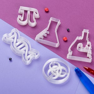 Puede incluir: Un conjunto de cinco cortadores de galletas de pl&aacute;stico blanco con forma de cadena de ADN, un vaso de precipitados, un microscopio, un s&iacute;mbolo pi y un s&iacute;mbolo de riesgo biol&oacute;gico. Los cortadores est&aacute;n sobre una superficie morada y morado claro con chispas rojas y negras.