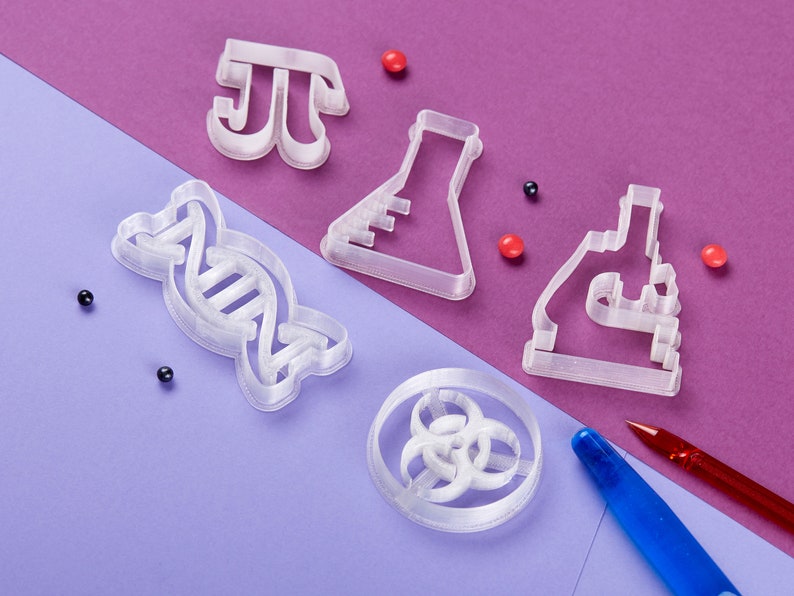 Puede incluir: Un conjunto de cinco cortadores de galletas de pl&aacute;stico blanco con forma de cadena de ADN, un vaso de precipitados, un microscopio, un s&iacute;mbolo pi y un s&iacute;mbolo de riesgo biol&oacute;gico. Los cortadores est&aacute;n sobre una superficie morada y morado claro con chispas rojas y negras.