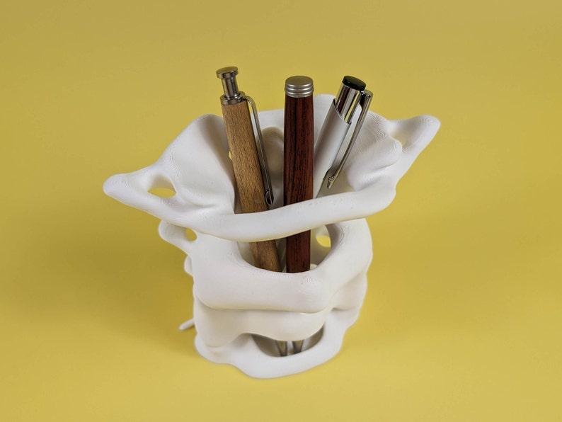 May include: White 3D printed pen holder shaped like a vertebrae, with three pens inside: a brown wooden pen, a dark red pen, and a silver pen.