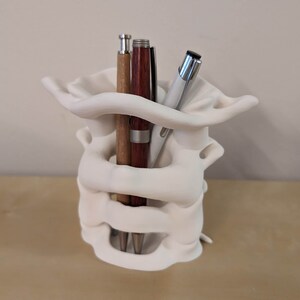 May include: White ceramic pen holder shaped like a human vertebra, with three pens inside. The pens are brown, silver, and black.