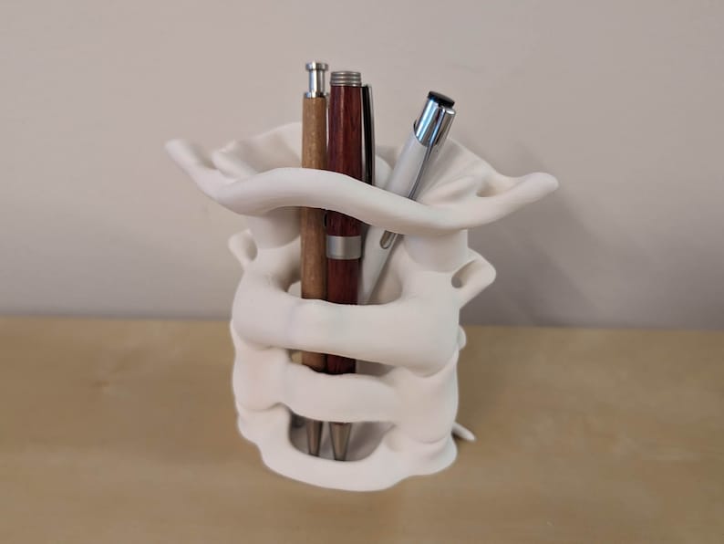 May include: White ceramic pen holder shaped like a human vertebra, with three pens inside. The pens are brown, silver, and black.
