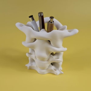 May include: White ceramic pen holder shaped like a human vertebra, with three pens inside.