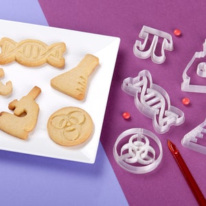 Puede incluir: Un plato blanco con galletas con tem&aacute;tica cient&iacute;fica, incluyendo una doble h&eacute;lice de ADN, un vaso de precipitados, un microscopio y un s&iacute;mbolo de riesgo biol&oacute;gico.  Junto al plato hay cortadores de galletas de pl&aacute;stico transparente en forma de doble h&eacute;lice de ADN, un vaso de precipitados, un microscopio, un s&iacute;mbolo de riesgo biol&oacute;gico y el s&iacute;mbolo matem&aacute;tico de pi.