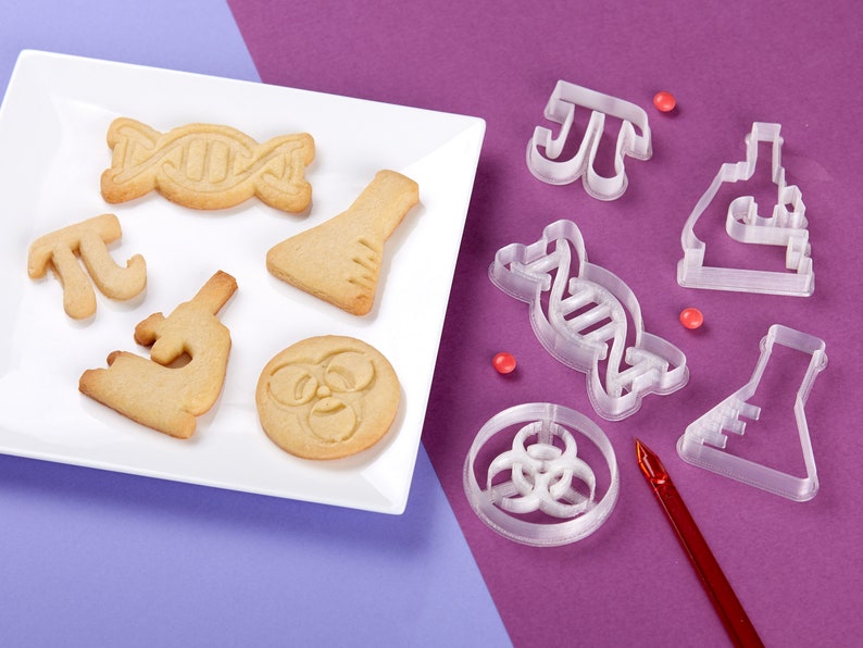 Puede incluir: Un plato blanco con galletas con tem&aacute;tica cient&iacute;fica, incluyendo una doble h&eacute;lice de ADN, un vaso de precipitados, un microscopio y un s&iacute;mbolo de riesgo biol&oacute;gico.  Junto al plato hay cortadores de galletas de pl&aacute;stico transparente en forma de doble h&eacute;lice de ADN, un vaso de precipitados, un microscopio, un s&iacute;mbolo de riesgo biol&oacute;gico y el s&iacute;mbolo matem&aacute;tico de pi.