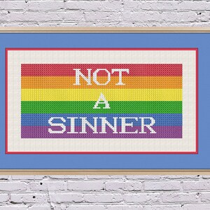 Peut inclure: Un motif de point de croix encadré représentant un drapeau arc-en-ciel avec le texte "NOT A SINNER" en blanc.