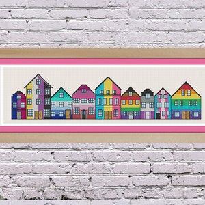 Puede incluir: Una obra de arte de punto de cruz enmarcada que representa una fila de casas coloridas con diferentes estilos arquitectónicos. Las casas están dispuestas en línea y presentan varios colores, incluidos amarillo, azul, verde, rojo y morado.