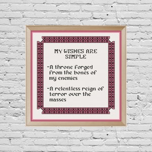 Könnte beinhalten: Ein gerahmtes Kreuzstich-Design mit einem rosa und schwarzen Rand. Der Text lautet "Meine Wünsche sind einfach - Ein Thron, geschmiedet aus den Knochen meiner Feinde - Eine unbarmherzige Herrschaft des Terrors über die Massen."