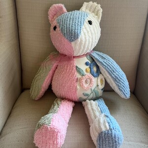 May include: A handmade plush bear with a patchwork design. The bear features pink, blue, white, and green sections. The bear has embroidered floral details on its chest and a pink ribbon around its neck. The bear is sitting on a neutral-colored chair.