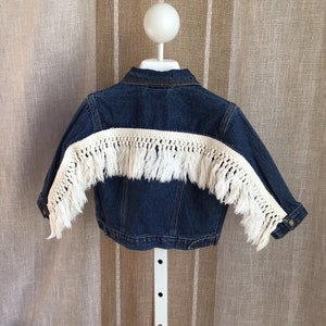 May include: A blue denim jacket with a white fringe detail across the back. The jacket is displayed on a white stand against a neutral background. The fringe adds a bohemian flair to the classic denim style.