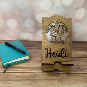 以下が含まれることがあります： カメの切り抜きと「Heidi」という名前が刻まれた木製の携帯電話スタンドです。