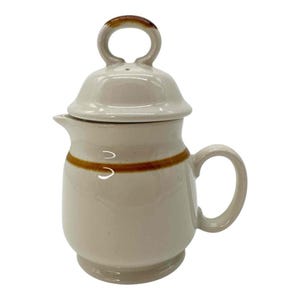 Puede incluir: Cafetera de cerámica color crema con tapa y asa. La cafetera tiene una franja marrón alrededor del centro y un asa curva. La tapa tiene un asa de bucle. El pico está en el lado izquierdo.