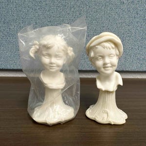 May include: Two off-white ceramic busts of children. One is in a clear plastic bag. The busts have detailed facial expressions and are mounted on decorative pedestals. The bust on the right is wearing a hat.