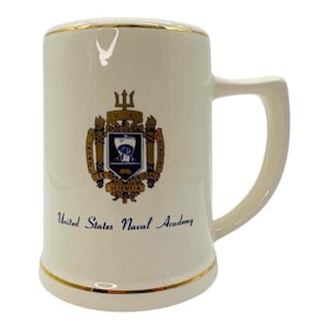 May include: Cream-colored ceramic mug with a gold rim and handle. The mug features the United States Naval Academy crest and the words "United States Naval Academy" in blue text.