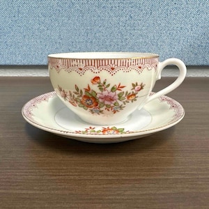 Puede incluir: Un juego de taza y platillo de té vintage. La taza y el platillo son blancos con un diseño floral en tonos de rosa, naranja y verde. El borde de la taza y el platillo está adornado con un patrón granate y dorado.