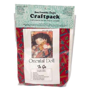May include: A craft kit for making an Oriental doll. The kit includes fabric, thread, and instructions. The doll is wearing a red and gold kimono with a white flower on her chest. The kit is packaged in a clear plastic bag with a red and white background.