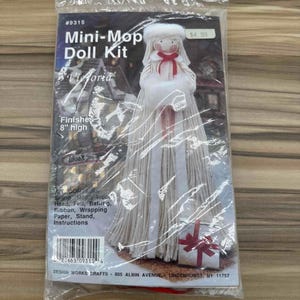 Puede incluir: Un kit de muñeca Mini-Mop, "Victoria", en un embalaje de plástico transparente. El kit incluye materiales para crear una muñeca de 20 cm de altura. La muñeca tiene una falda de fregona blanca, un cuello de piel blanco y una cinta roja.
