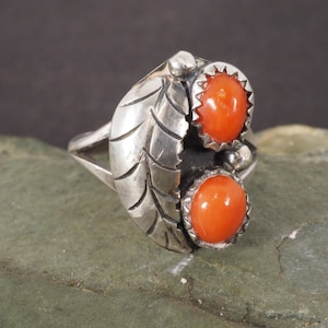 May include: A silver ring featuring a leaf design with two oval, orange-red gemstones. The gemstones are set in a scalloped bezel. The ring is on a green and gray surface.