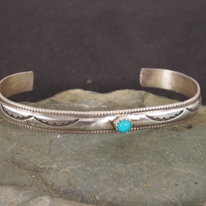 May include: A silver cuff bracelet featuring a thin, polished band with a beaded edge and engraved crescent patterns. A single, small, round, bright turquoise stone is set in the center of the metal band, which has an open back design.