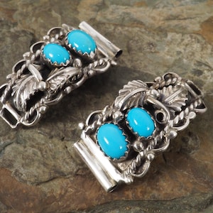 May include: A pair of silver bracelet clasps, each with three oval turquoise stones. The clasps have intricate silver detailing, including leaves and vines. The bright blue turquoise stones are set in the silver. The clasps are displayed on a textured, stone surface.