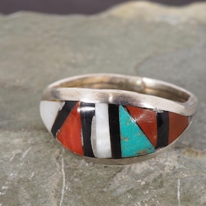 May include: Silver ring with a colourful inlaid design. The ring features a pattern of white, black, red, and turquoise geometric shapes. The ring is set against a neutral background.