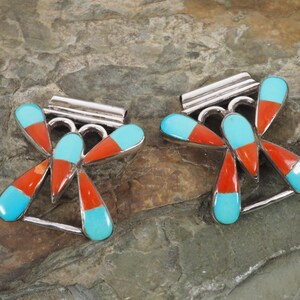 May include: A pair of silver-toned earrings with a dragonfly design. Each earring features a central silver bar with a series of curved accents. The dragonfly wings are composed of turquoise and orange-red teardrop-shaped stones.