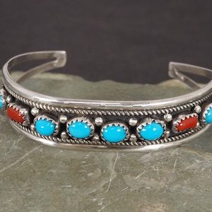 May include: A silver cuff bracelet featuring a row of alternating oval turquoise and red coral cabochons set in serrated bezels, accented by twisted wire rope detailing and small silver spheres along the center band of the metal cuff.