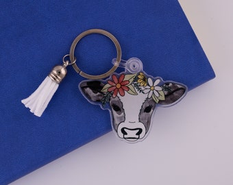 May include: A white cow keychain with a floral crown, black and white spots, and a white tassel. The keychain is made of clear acrylic and has a silver metal ring.