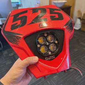 Può includere: Un faro per moto rosso con il numero "525" in nero. Il faro ha quattro luci rotonde e un inserto in schiuma nero. I fili si estendono dalla parte inferiore.