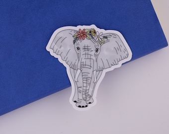 May include: A white elephant sticker with a flower crown on its head. The flowers are pink, white, and yellow. The elephant is outlined in black.