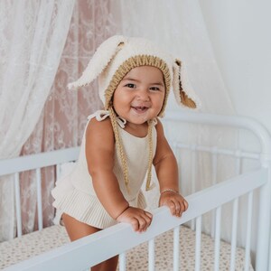 May include: A baby wearing a white and brown bunny hat is standing in a white cot with a floral patterned sheet. The baby is smiling and looking at the camera.