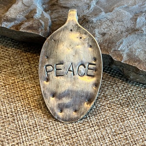 May include: A silver spoon pendant with the word "PEACE" embossed on it. The pendant is sitting on a brown burlap surface.