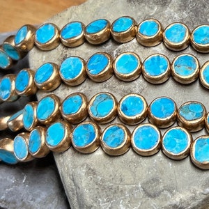 May include: Turquoise blue round beads with gold accents. The beads are arranged in rows on a gray stone surface.