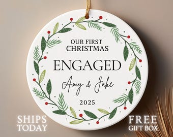 May include: A white ceramic Christmas ornament with the text "OUR FIRST CHRISTMAS ENGAGED Amy & Jake 2025". The ornament is round and decorated with a green and red floral wreath and a gold string.