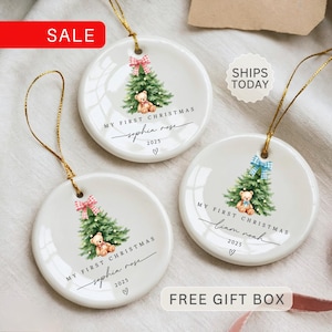 May include: White ceramic Christmas ornaments with gold string. Each ornament features a watercolor Christmas tree, teddy bear, and the text "MY FIRST CHRISTMAS" with a name and the year 2025. The ornaments have red and blue bows.