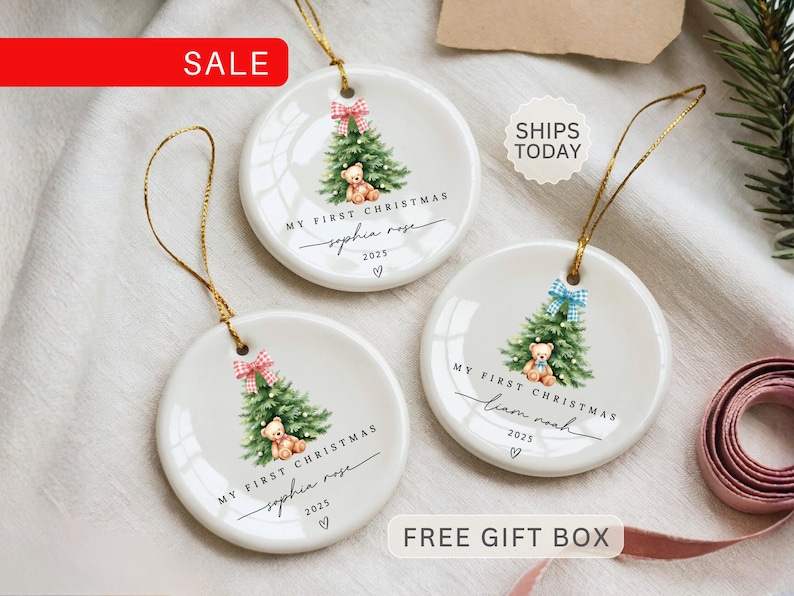 May include: White ceramic Christmas ornaments with gold string. Each ornament features a watercolor Christmas tree, teddy bear, and the text "MY FIRST CHRISTMAS" with a name and the year 2025. The ornaments have red and blue bows.