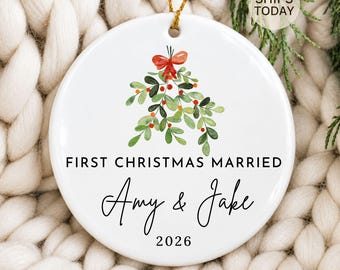 Könnte beinhalten: Ein rundes, weißes Keramik-Weihnachtsornament mit einer Aquarell-Illustration von Misteln und einer roten Schleife. Der Text lautet FIRST CHRISTMAS MARRIED, Amy & Jake und 2026 in schwarzer Schrift. Ein goldener Faden dient zum Aufhängen.